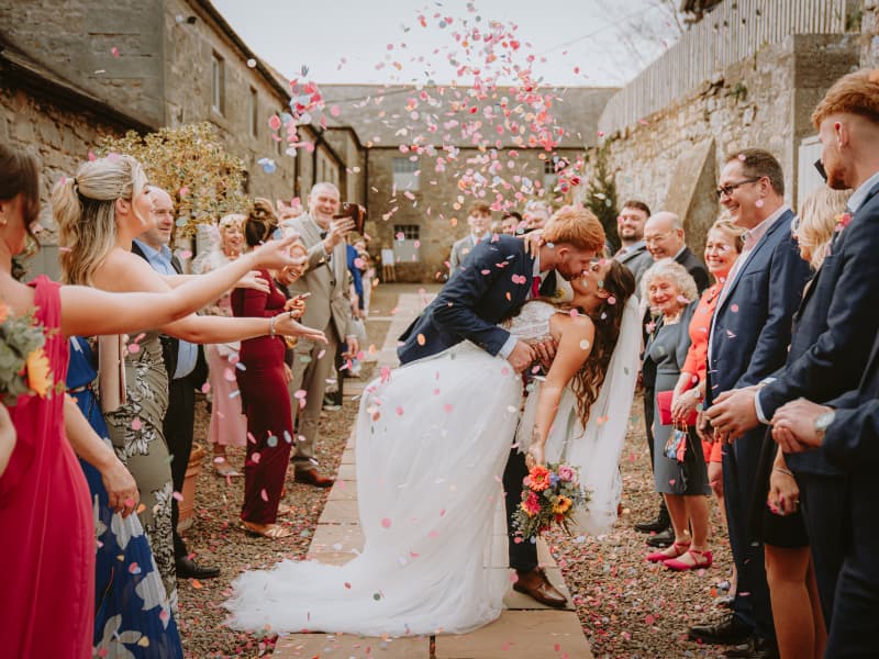 ewlywed couple share a kiss as guests throw pink and white confetti around them in a stone courtyard, creating a celebratory wedding exit scene.